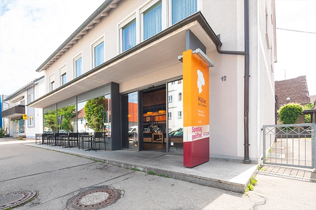 Außenansicht der Filiale in Grossaitingen. Zu sehen ist die Häuserfassade, die Außenbestuhlung mit Sitzbereich sowie das orangene Eingangschild der Bäckerei Müller am Eingang.