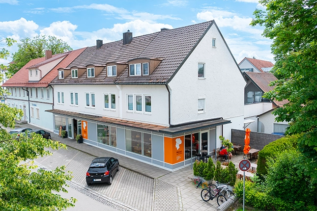 Außenansicht der Filiale in Klosterlechfeld. Zu sehen ist die Häuserfassade mit großer Glasfront durch die man in die Filiale blicken kann. Vornedran sind die Parkplätze der Bäckerei Müller. Rechts von der Filiale ist der Außenbereich mit Stühlen tischen sowie Sonnenschirmen zu sehen.