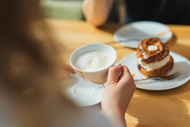 Kunden trinkt einen Cappuccino der Bäckerei Müller. Im Hintergrund ist ein Kirschringmit fruchtiger Kirschfüllung und Streuseln zu sehen.