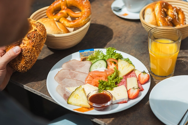 Schlemmerfrühstück, bestehend aus Schinken, Salami, Käse, Lachs, Butter, Marmelade, ein weiches Ei und einem Brotkörbchen. Ebenso zu sehen ist ein Glas Orangensaft.