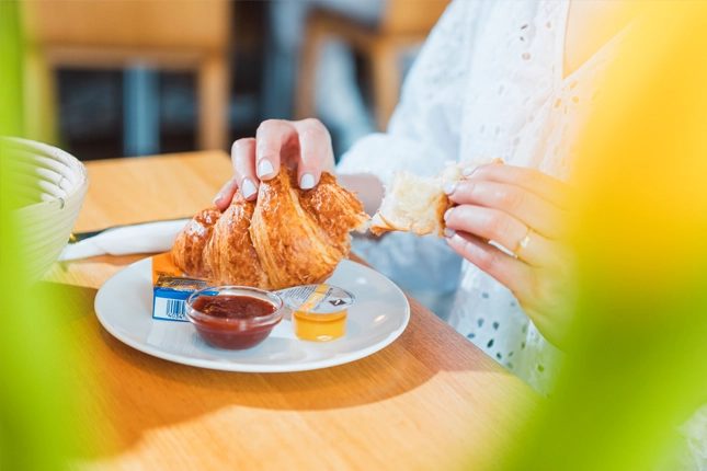 Ein weiblicher Gast reißt ein frisches Croissant auf, um es mit Butter, Marmelade und/oder Honig zu essen. Das Die Komponenten bilden das französische Frühstück der Bäckerei Müller.