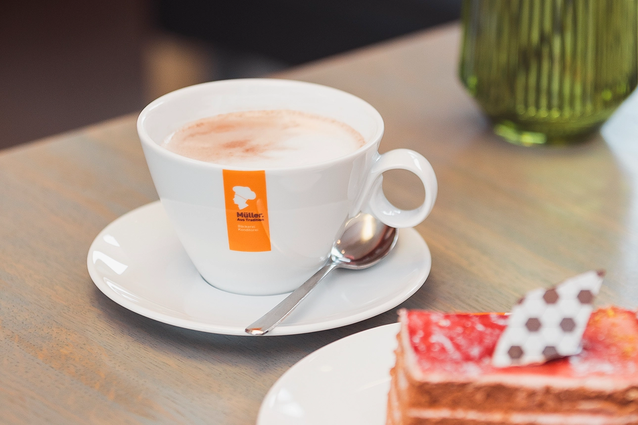 Tasse heiße Schokolade mit Löffel auf einer Untertasse. Im Vordergrund zeichnet sich ein Stück Kuchen ab.