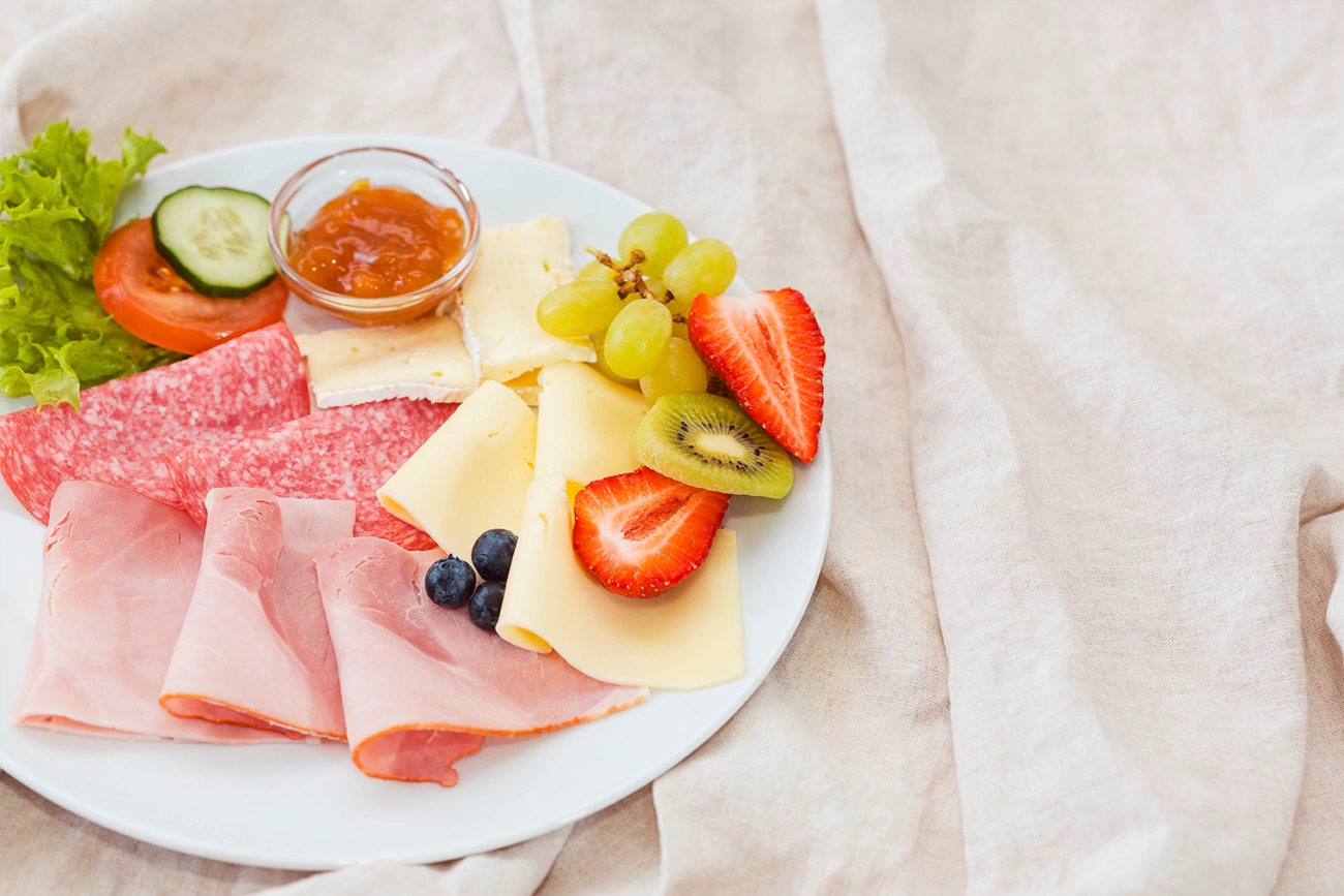 Frühstücksteller des klassischen Frühstücks mit Schinken, Salami, Käse, Butter, Marmelade, Salat, Gurke, Tomate, Blaubeeren, Erdbeeren und zwei Semmeln auf Leinenstoff drapiert