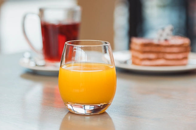 Glas frischer Orangensaft. Im Hintergrund ist eine Tasse Tee sowie ein geschichteter Kuchen auf dem Tisch zu erkennen.