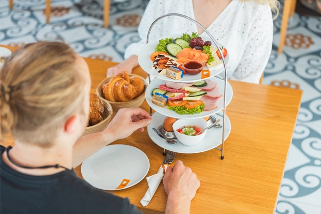 Das Gute-Laune-Frühstück auf einer Etagere serviert. Zu sehen sind Wurst- und Käsevariationen, Lachs, zwei weiche Eier, zwei kleine Früchtejoghurts, Marmelade, Butter und zwei Brotkörbchen mit Semmeln und Croissants. Der männliche Gast greift nach einem Löffel, um sein Frühstücksei zu essen.