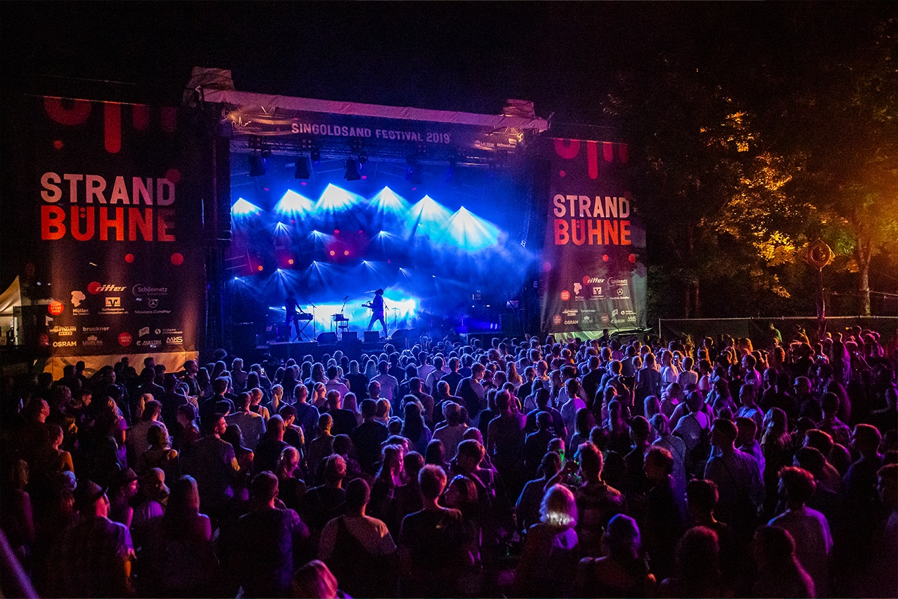 Impression des Singoldsand Festivals 2019. Neugierige und musikbegeisterte Gäste stehen versammelt vor der mit blauem Bühnenlich gefluteten Bühne und hören dem Sänger gespannt bei seiner Performance zu