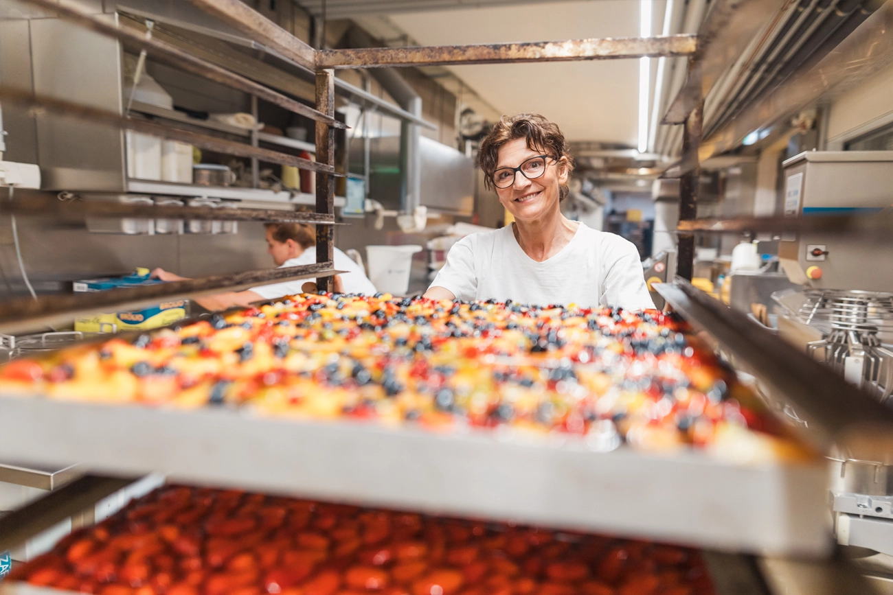 Mitarbeiterin, die riesige Kuchenbleche im Edelstahltrolley vor sich her schiebt. Unter anderem ist ein bunter Fruchtkuchen mit diversen Früchten sowie ein Erdbeerkuchen zu erkennen.