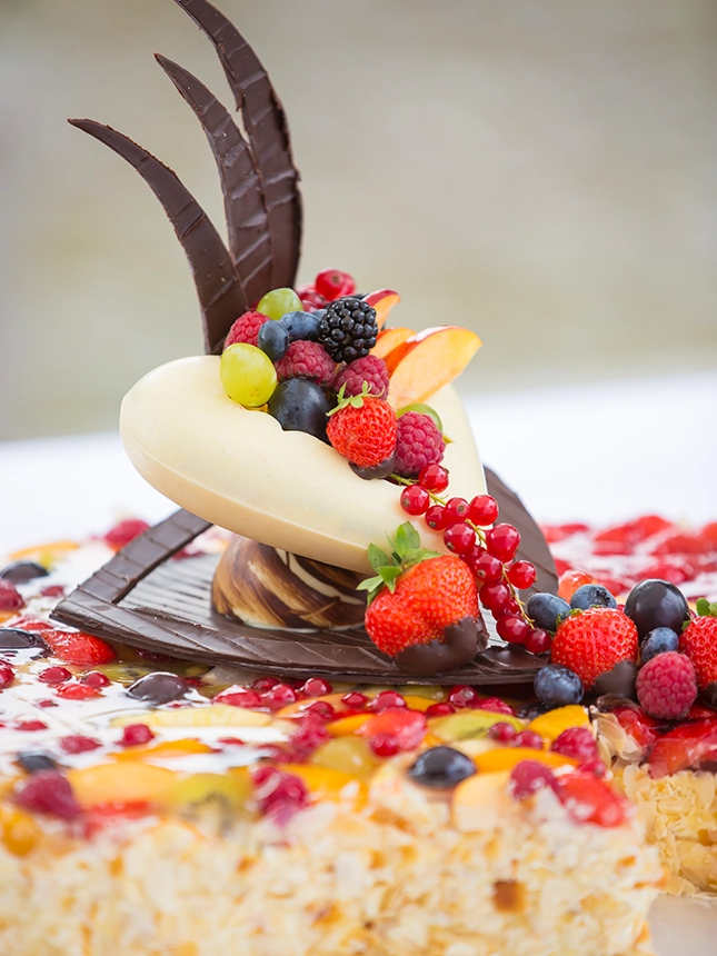 Detailaufnahme eines Toppers einer Hochzeitstorte in doppelter Herzform auf Früchtebasis. Das Herz ist aus weißer Schokolade geformt, mit einem Fächer aus dunkler Schokolade verziert und mit diversen frischen Beeren und Früchten gefüllt.