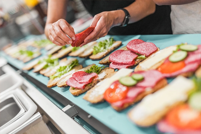 Nahaufnahme von zwei Händen, die eine Reihe an Semmeln und Seelen für die Snacktheke der Bäckerei Müller belegt.