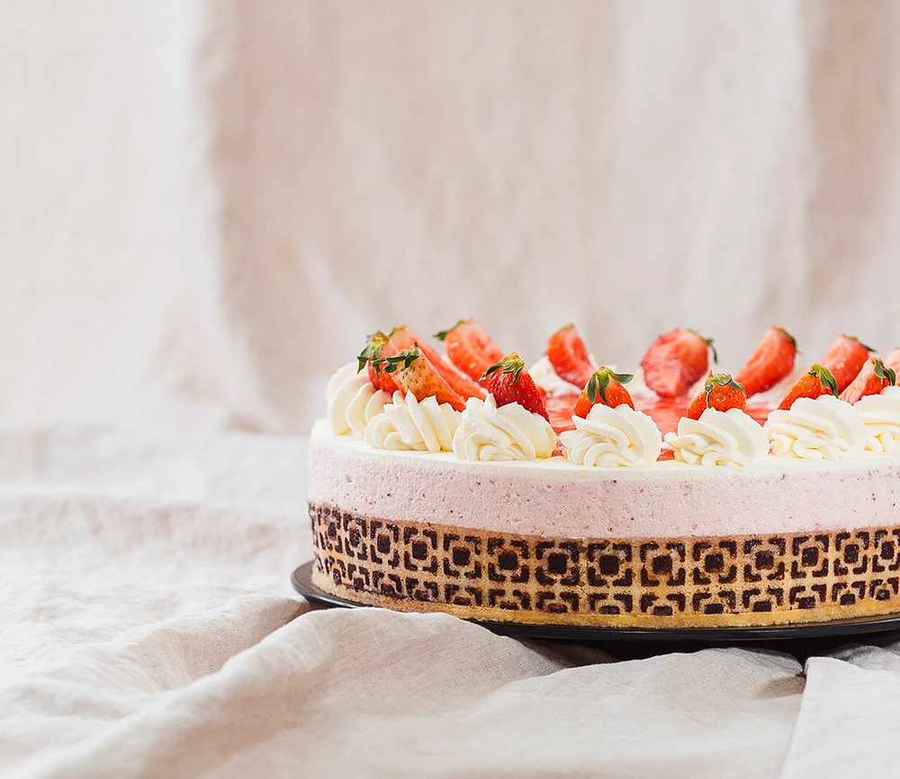 Detailaufnahme einer Erdbeertorte mit kunstvollem Rand und Sahnehäubchenverzierung und frischen Erdbeeren. Die Torte wurde auf einem schwarzen Teller auf Leinenstoff drapiert.