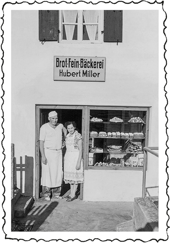 Bild von Großvater Hubert Miller und seiner Frau. Auf den Werbeschild über dem Bäckereieingang steht "Brot·Fein·Bäckerei Hubert Miller". Damals wurde der Nachname der Familie Müller fälschlicherweise mit einem "i" anstelle eines "ü’s" abgedruckt.